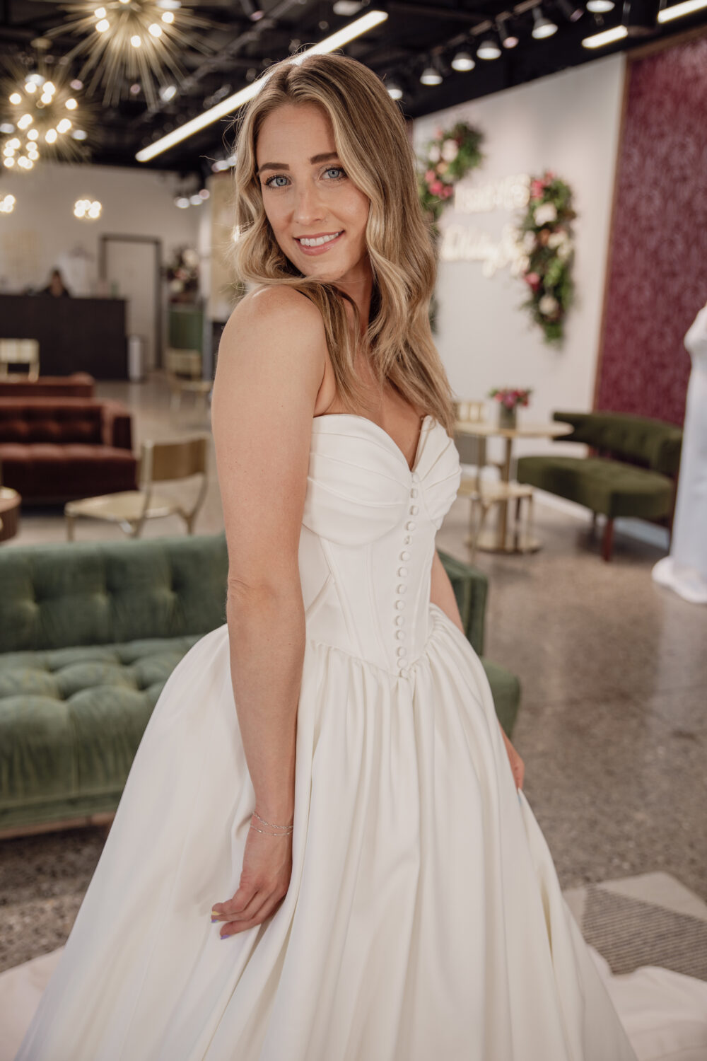 Bride smiling in a strapless white wedding dress with a buttoned corset-style bodice inside a bridal boutique.