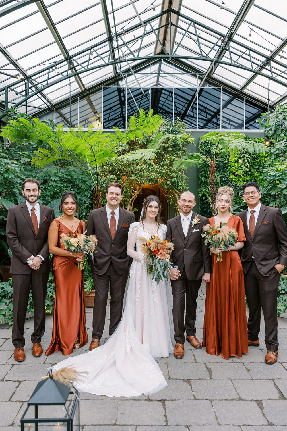A wedding party poses inside a glass-roofed conservatory, with the bride in white, attendants in rust-colored dresses, and men in dark suits.