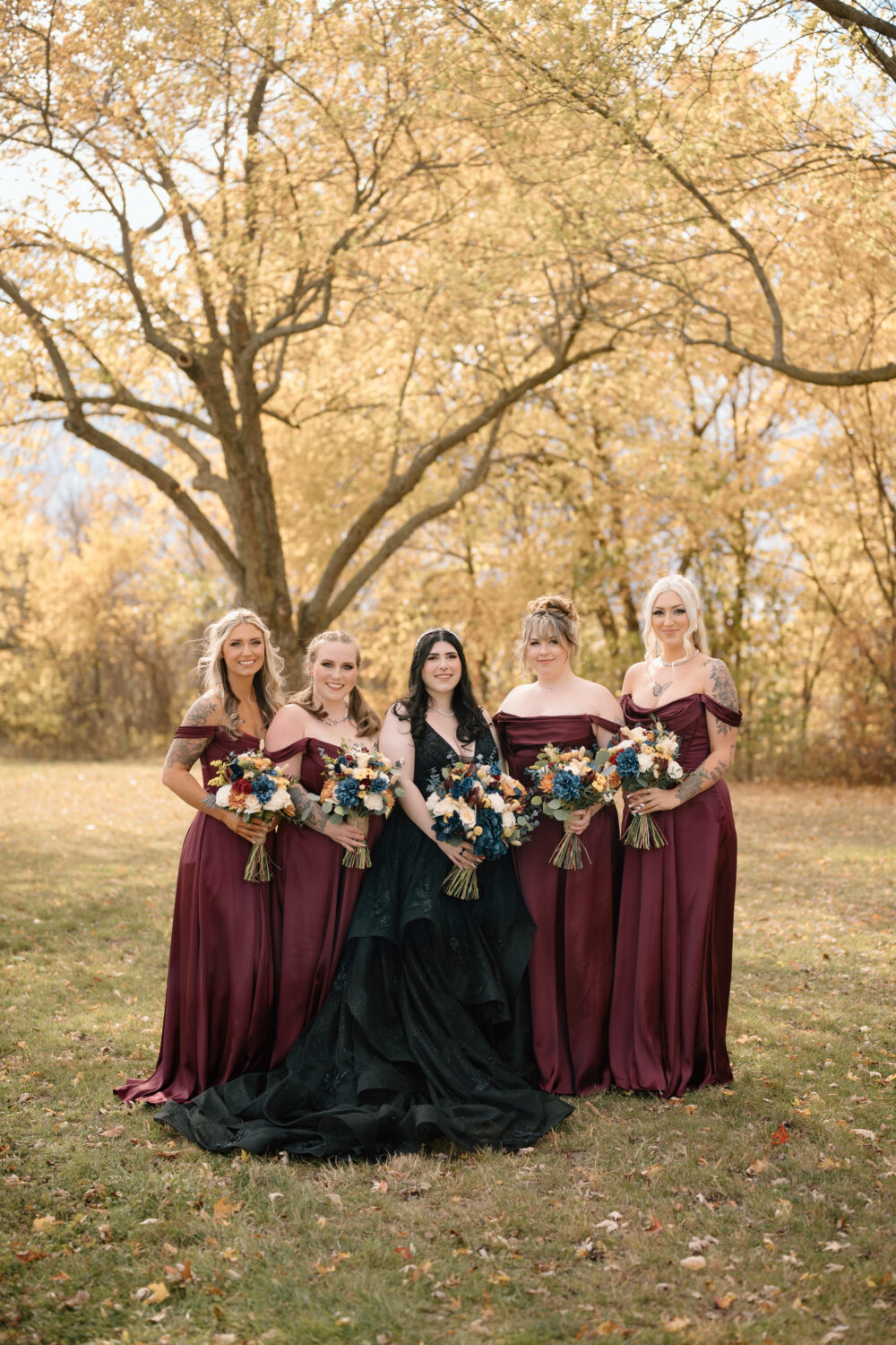 A bride in a black wedding gown stands outdoors with four bridesmaids in burgundy dresses beneath a large tree with golden autumn leaves.