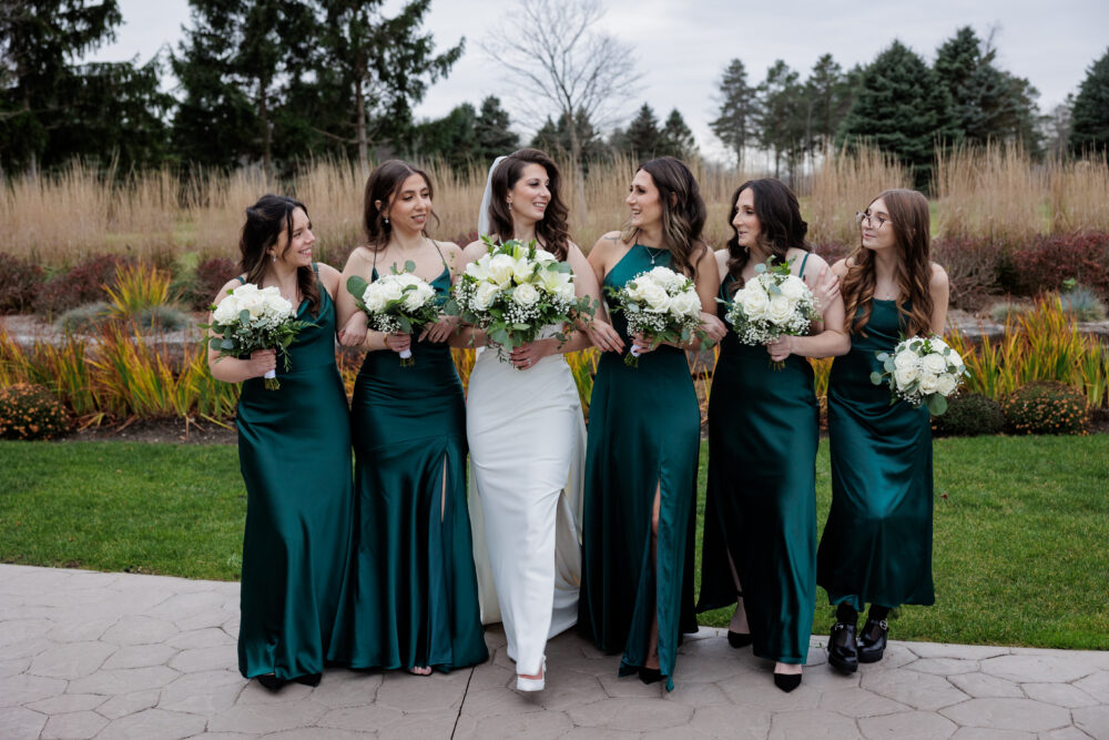 A bride in a fitted white wedding dress walks outdoors with five bridesmaids wearing floor-length dark green dresses and carrying white bouquets.
