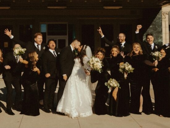 A large wedding party dressed primarily in black formalwear poses together outside a building, with the bride in a white wedding gown at the center.
