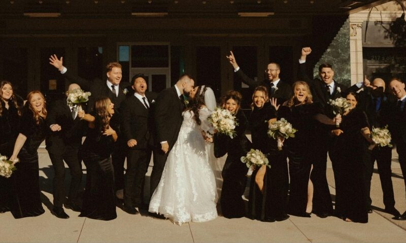 A large wedding party dressed primarily in black formalwear poses together outside a building, with the bride in a white wedding gown at the center.
