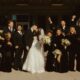 A large wedding party dressed primarily in black formalwear poses together outside a building, with the bride in a white wedding gown at the center.