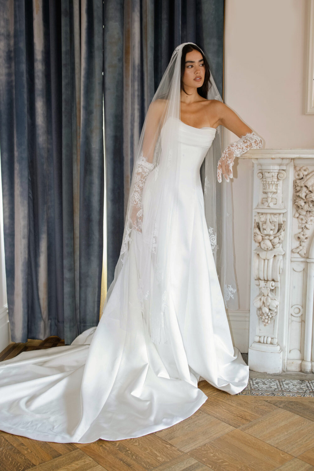 la premiere bride wearing lace gloves and a veil, leaning on a marble fireplace.