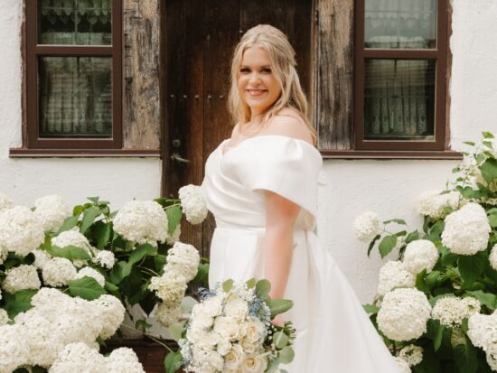 A bride in an off-the-shoulder wedding gown holds a white bouquet beside blooming white hydrangeas and a rustic wooden doorway.