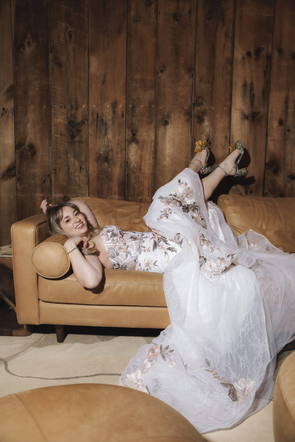 A woman reclines on a tan leather sofa wearing a flowing white gown with colorful floral details and a voluminous skirt.