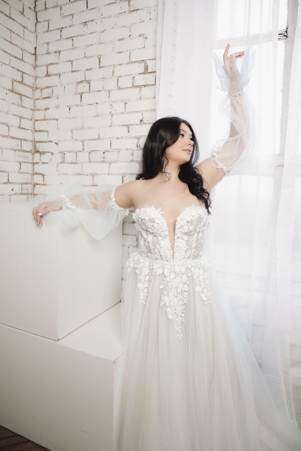 Bride in a white wedding gown with sheer off-the-shoulder sleeves posing beside a bright window and white brick wall.