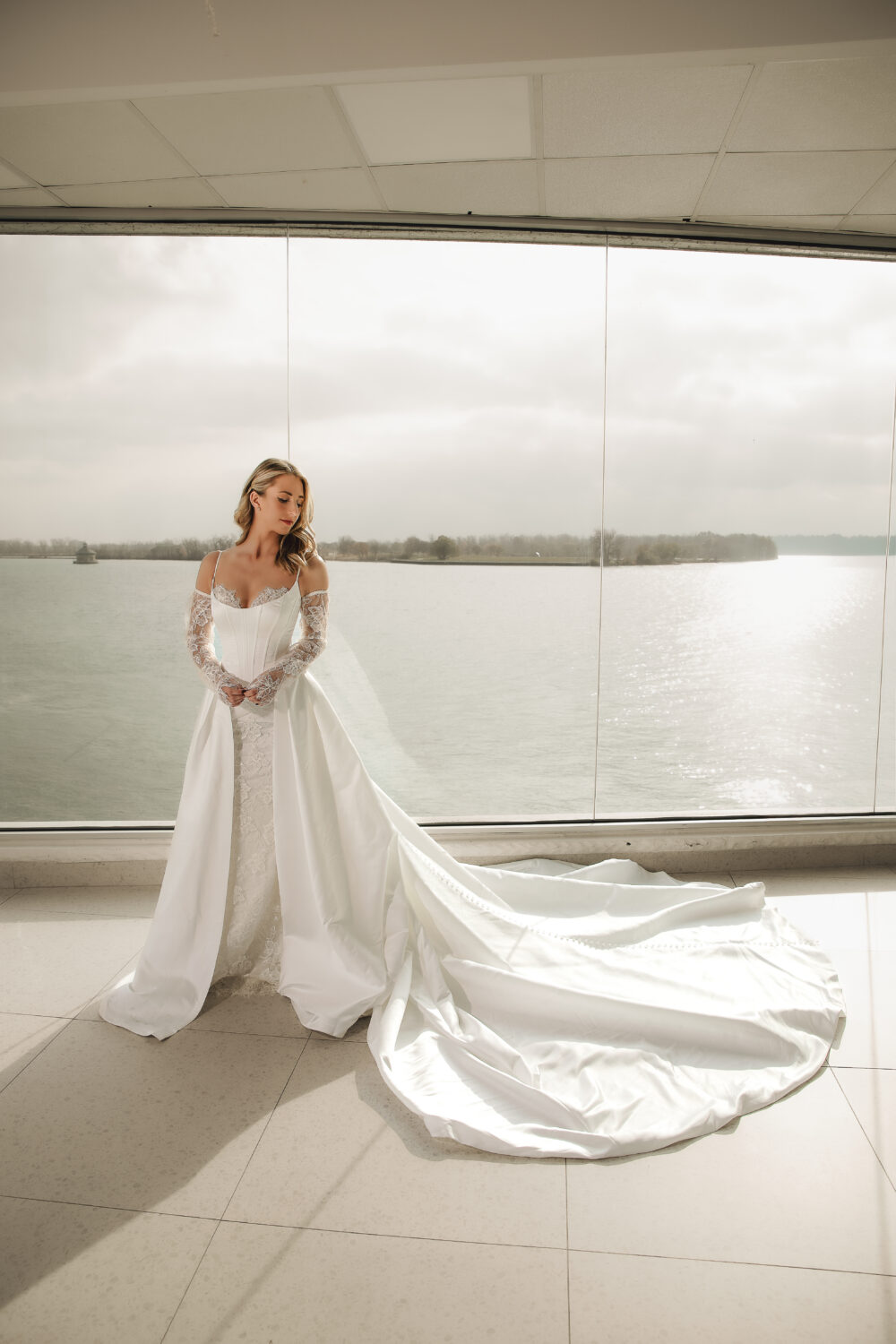 Bride in a white wedding dress with lace sleeves and a long flowing overskirt and train standing before floor-to-ceiling windows overlooking the water.