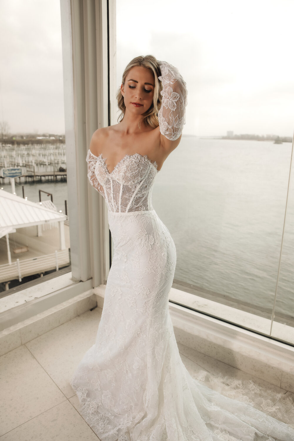 Bride in a fitted lace wedding gown with sheer lace sleeves posing beside large windows overlooking the water.