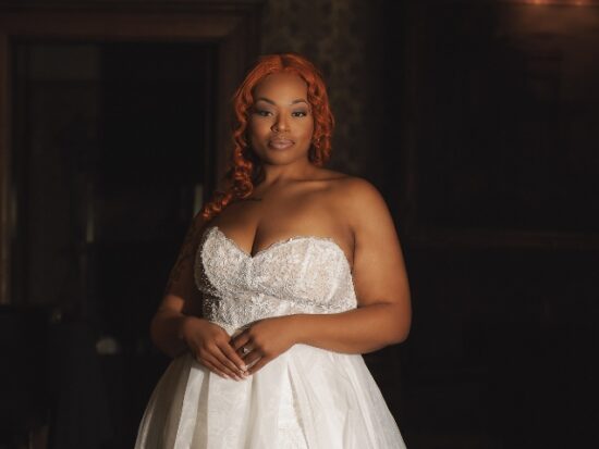 Bride with red hair wearing a strapless white wedding gown with a beaded bodice and flowing skirt in a dark wood-paneled doorway.