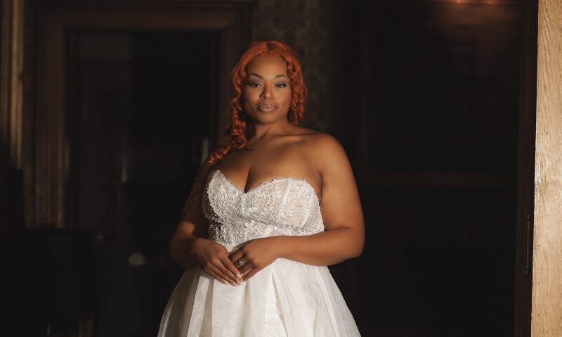 Bride with red hair wearing a strapless white wedding gown with a beaded bodice and flowing skirt in a dark wood-paneled doorway.