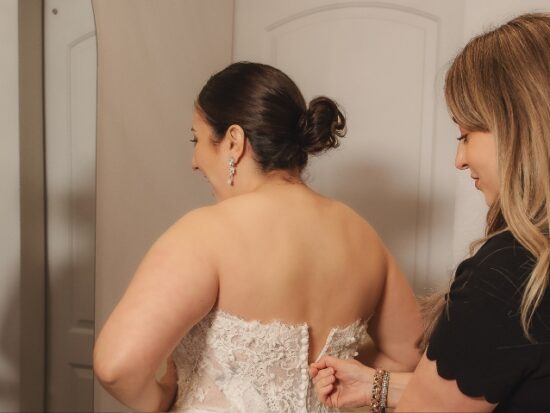 A woman in a black dress fastens the buttoned back of a lace wedding gown during a fitting.