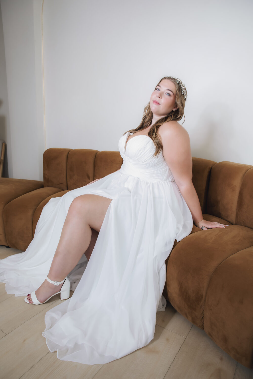 Bride seated on a brown sofa in a white wedding gown with a ruched sweetheart bodice, flowing skirt, and jeweled headband.