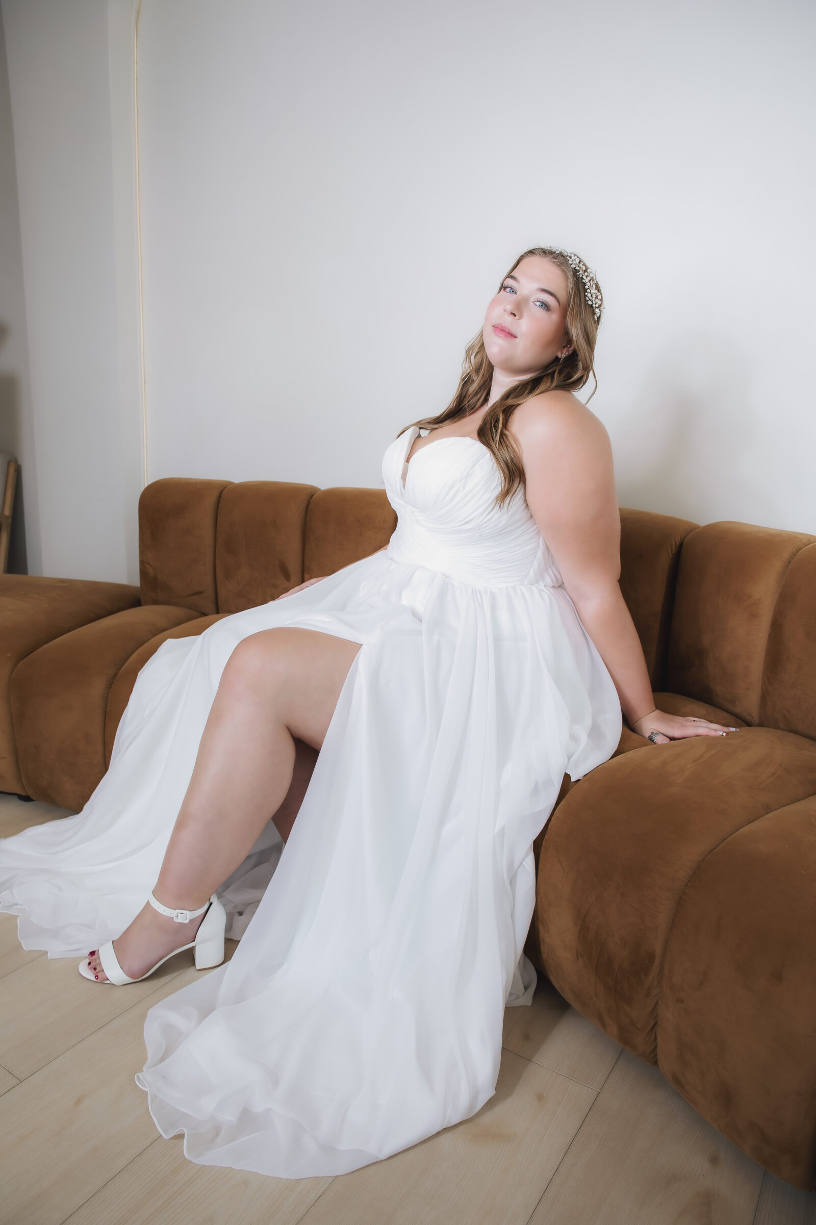 Bride seated on a brown sofa in a white wedding gown with a ruched sweetheart bodice, flowing skirt, and jeweled headband.