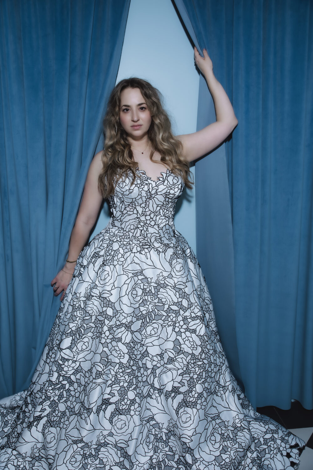 Bride posing between blue curtains in a strapless black-and-white floral ball gown with a full skirt.