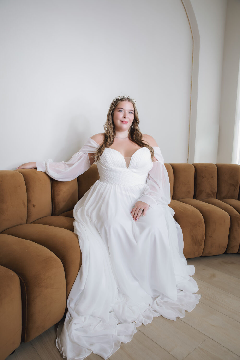 Bride seated on a brown sofa in a white wedding gown with a sweetheart neckline and sheer off-the-shoulder sleeves.