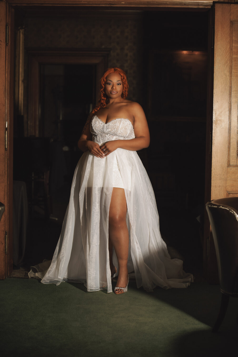 Bride with red hair wearing a strapless white wedding gown with an embellished bodice, high slit, and sheer overskirt in a dark doorway.