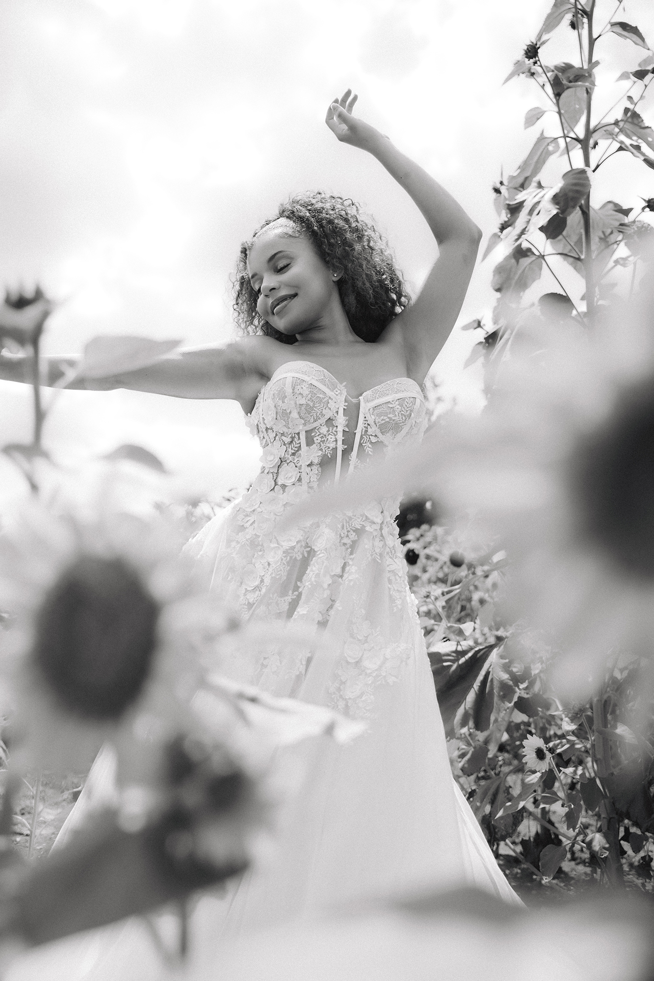 Bride in a white wedding gown posing outdoors among flowers in a black-and-white portrait.
