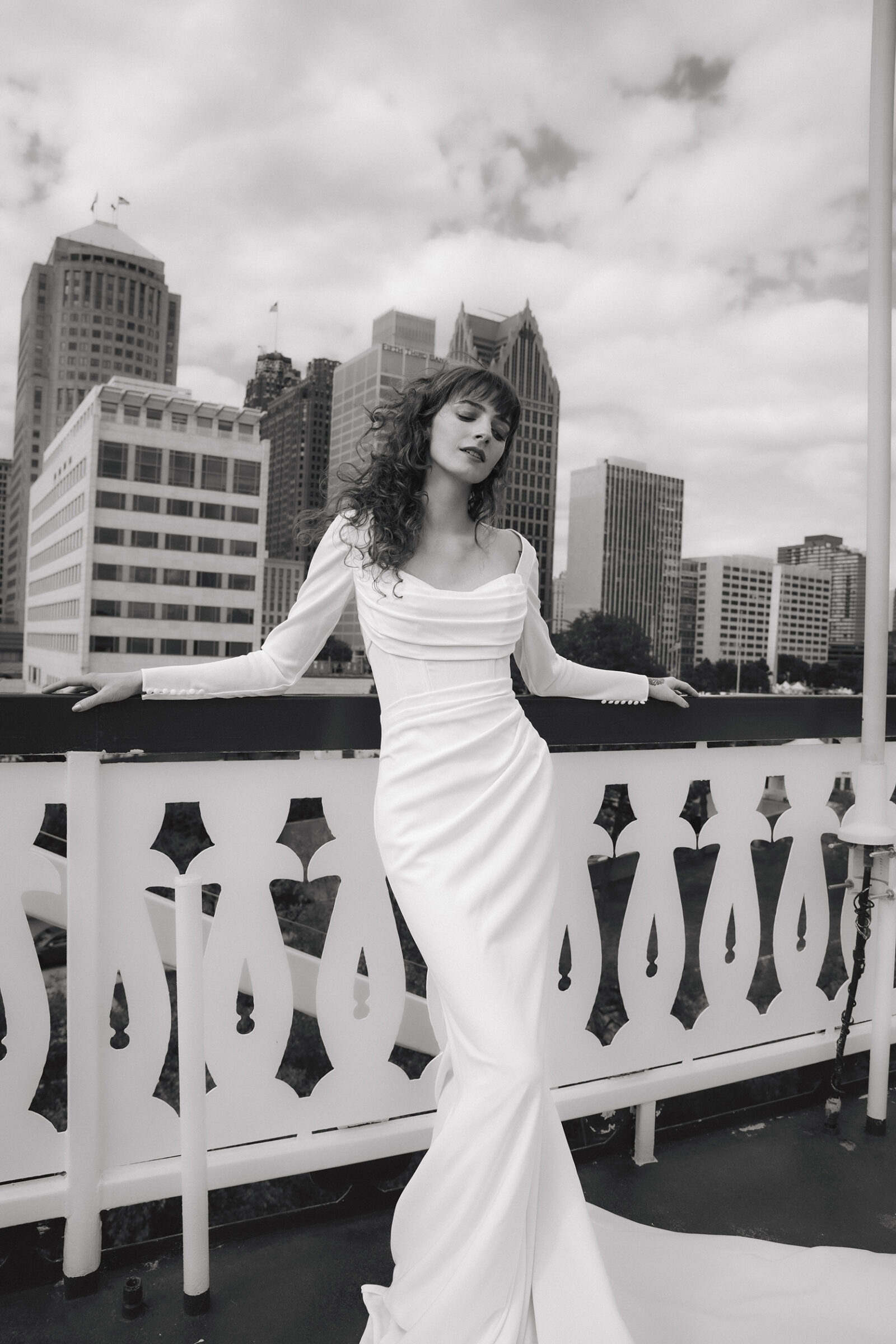 Bride in a fitted white wedding dress posing on a balcony with a city skyline in the background.