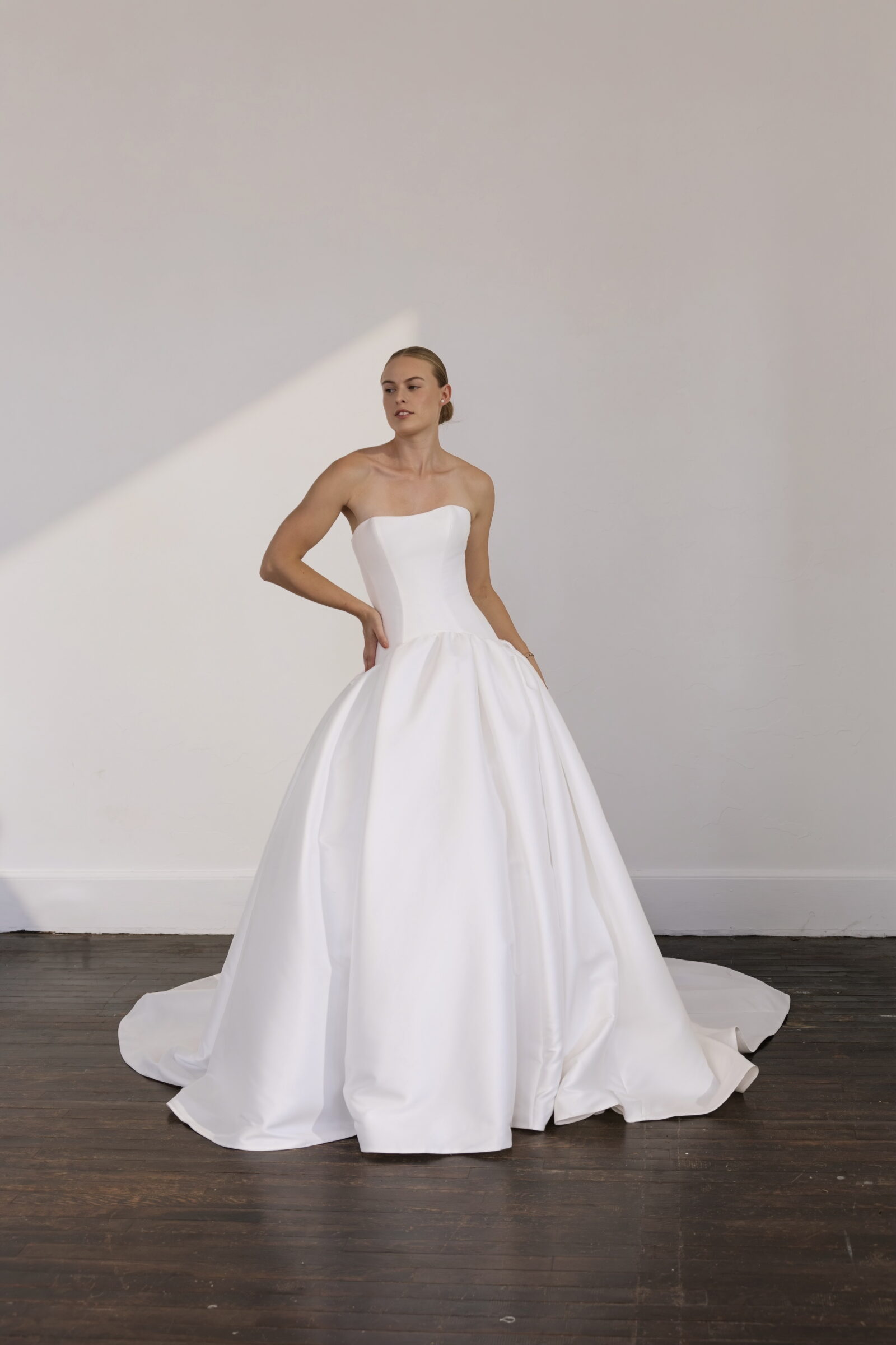 Bride wearing a strapless white satin ball gown with a fitted bodice and full skirt, posing in a minimalist studio.