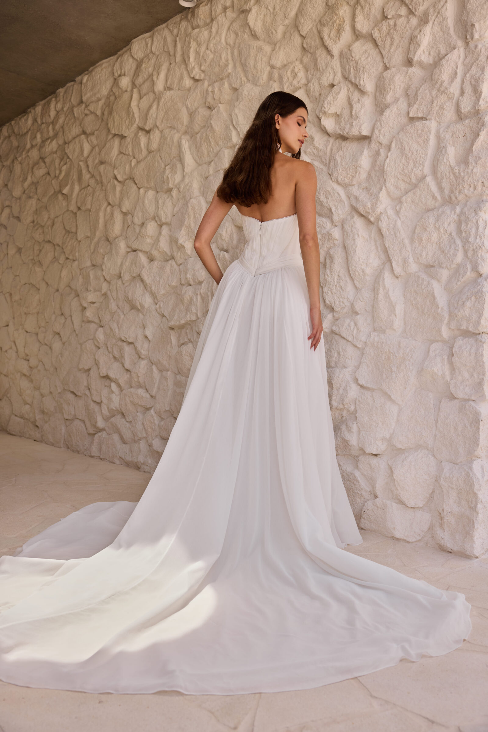 Bride wearing a strapless white wedding gown with a long flowing train, standing against a textured stone wall.