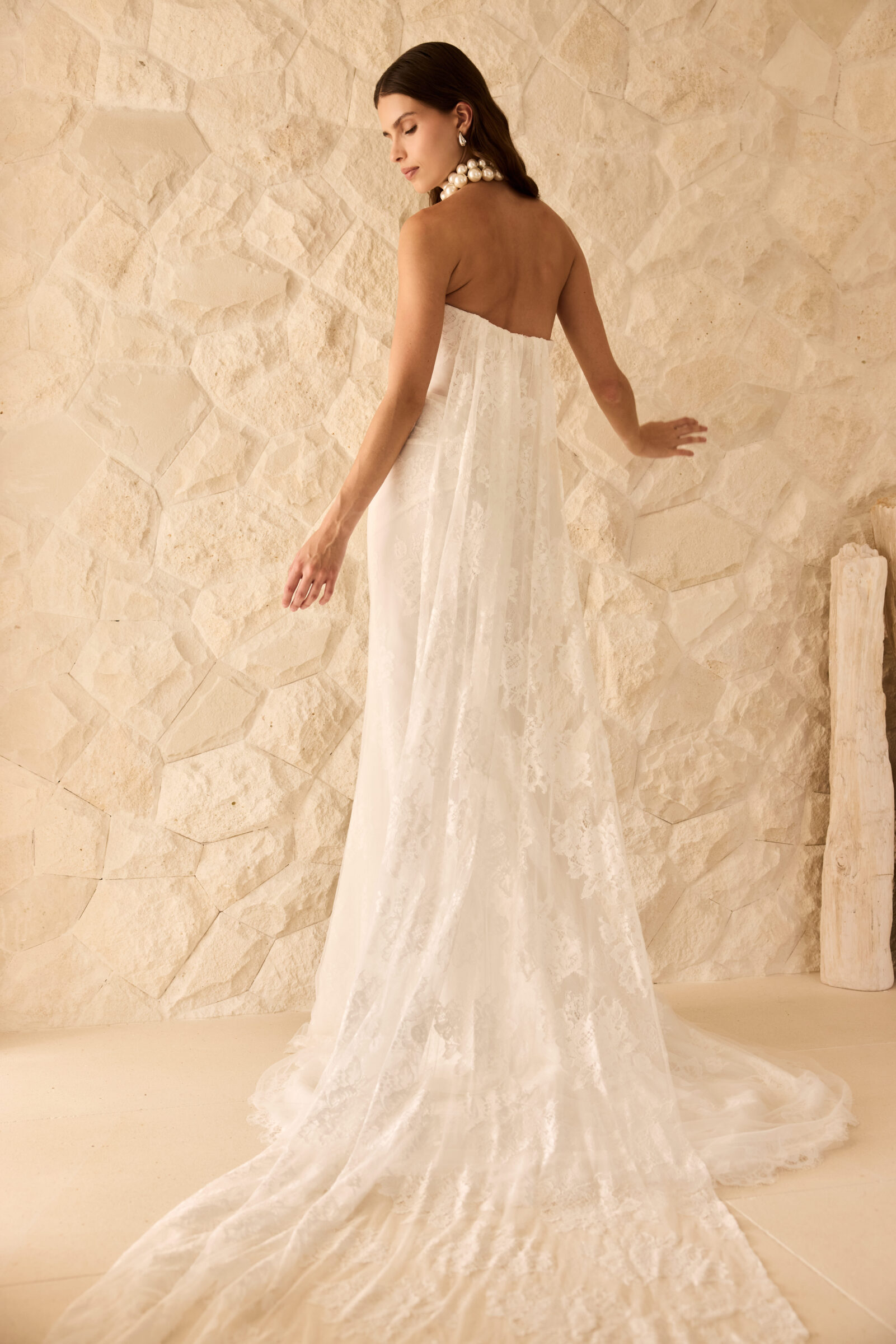 Bride wearing a strapless fitted white wedding gown with a long train, posing against a stone wall.