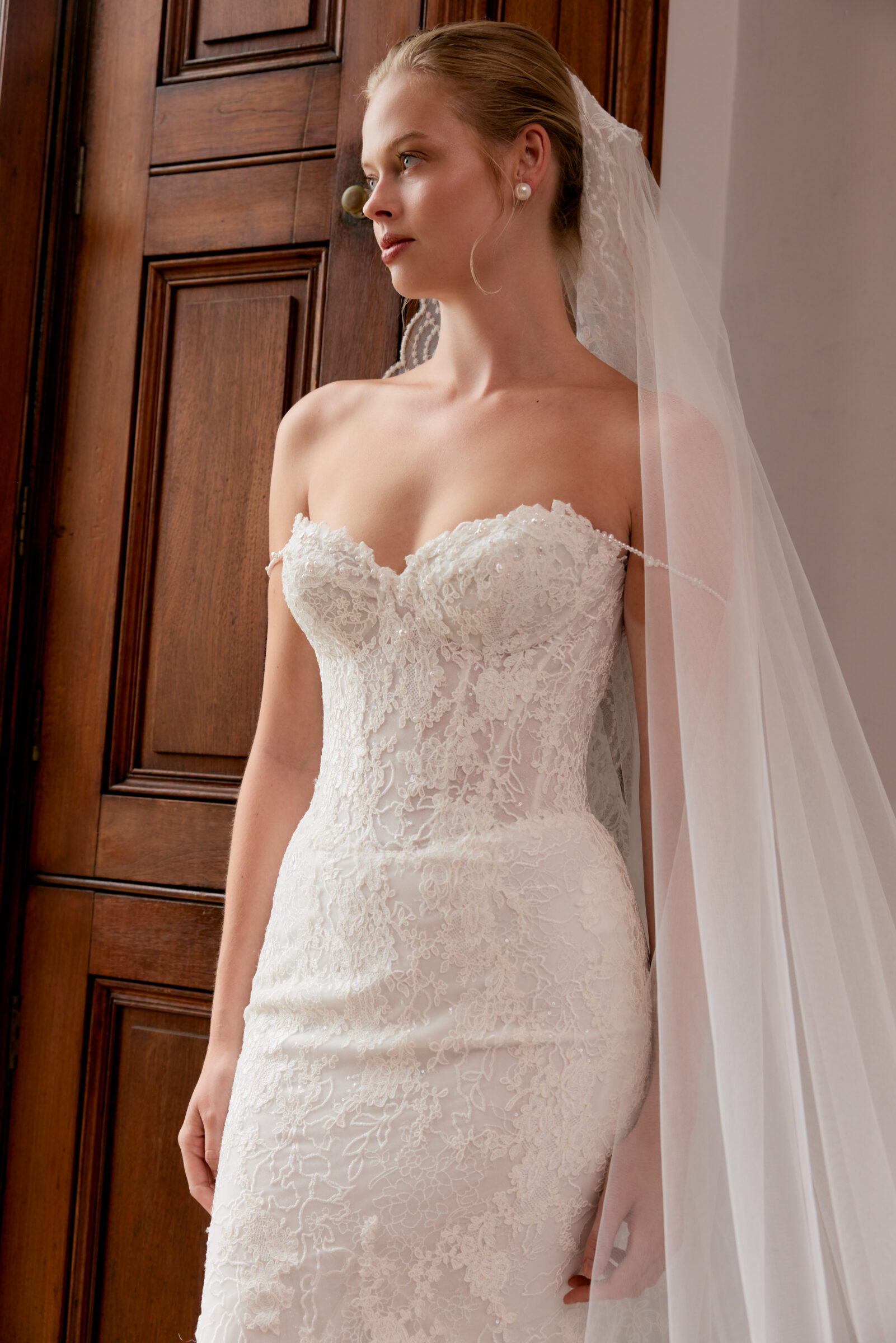 Close-up of a bride in a strapless lace wedding gown with a sweetheart neckline and long veil, standing beside a wooden door.