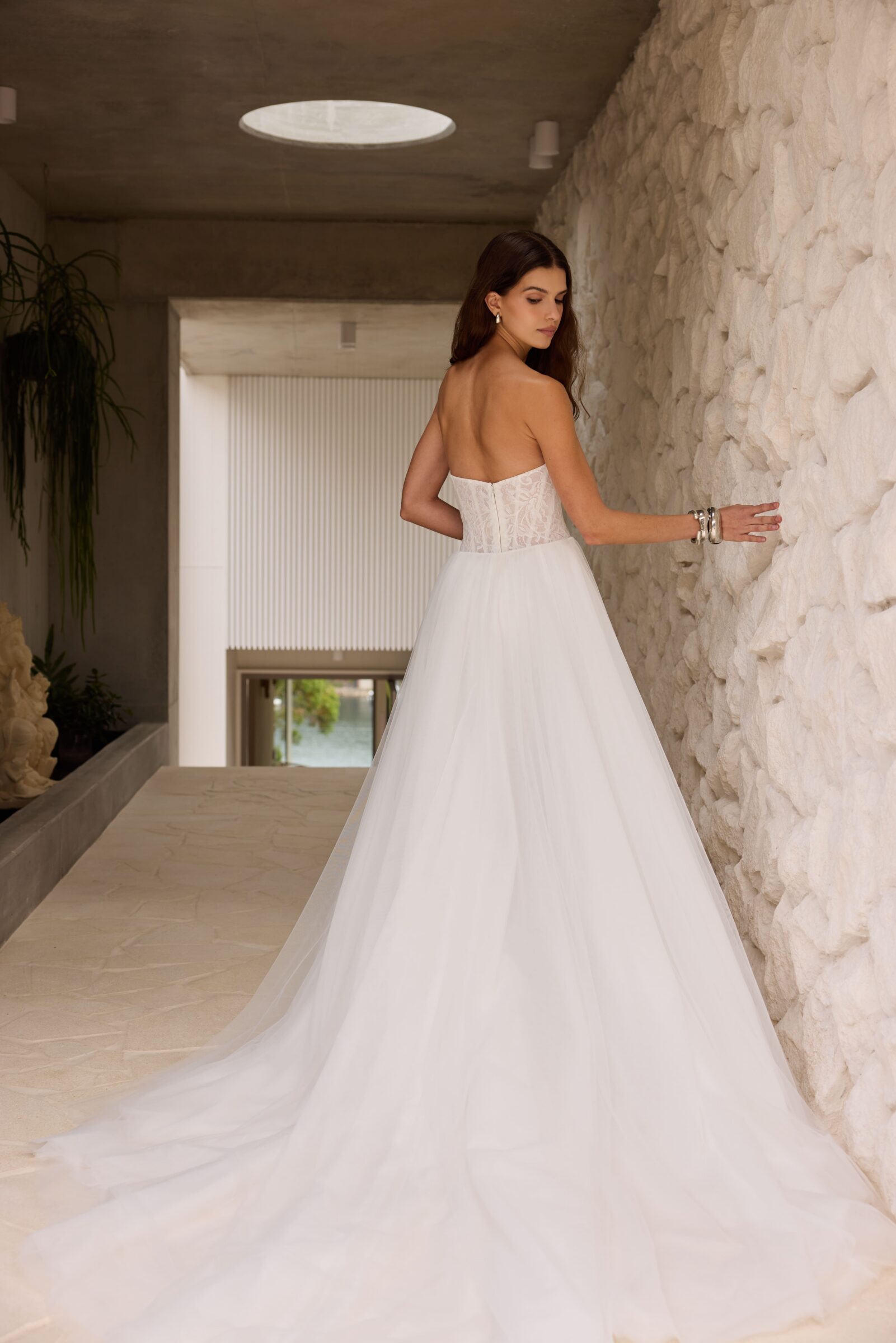 Bride in a strapless white wedding gown with a long flowing train, standing in a stone hallway.