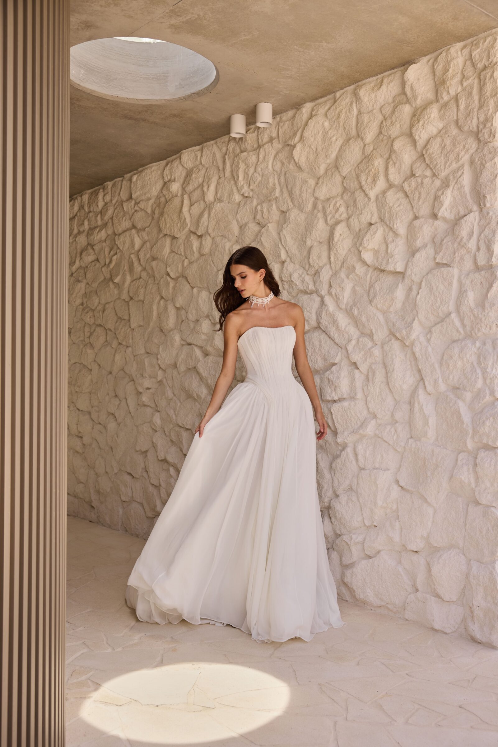 Bride wearing a strapless white wedding gown with off-the-shoulder draped sleeves and a pearl necklace, posing outdoors.