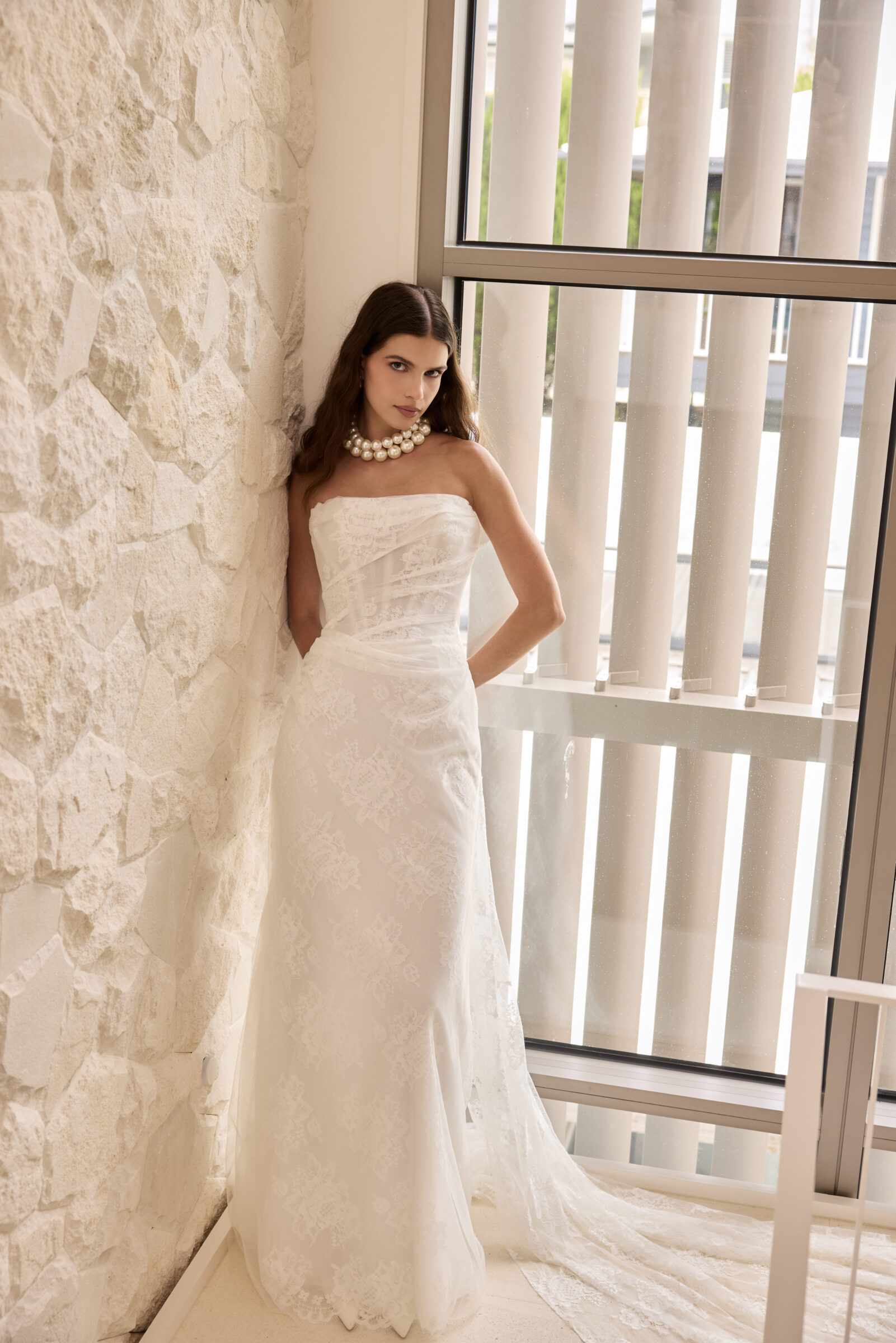 Bride in a strapless fitted white wedding gown, standing beside a large window in a stone interior.