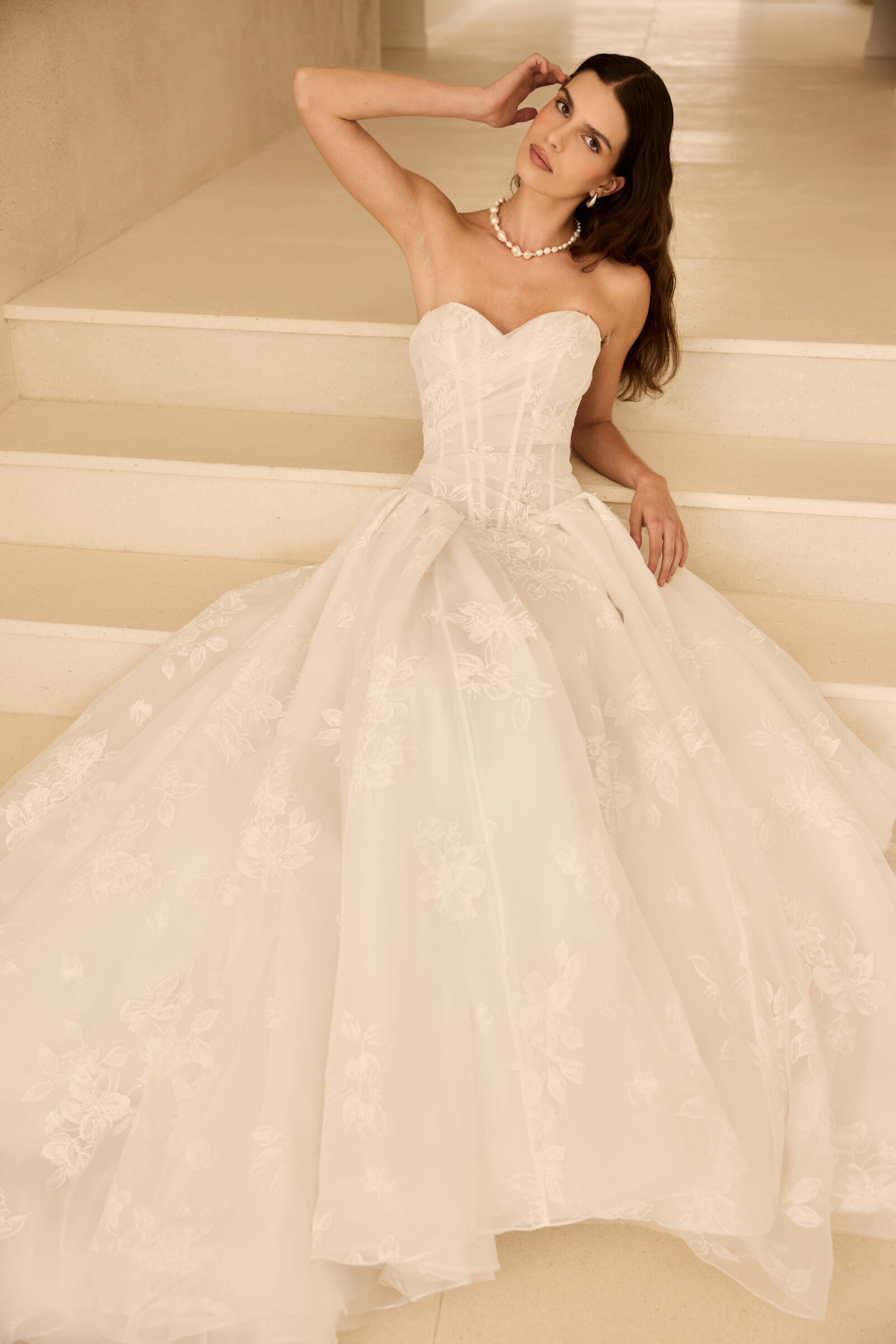 Bride wearing a strapless white wedding gown with a full, floral-embellished skirt, posing on a staircase.