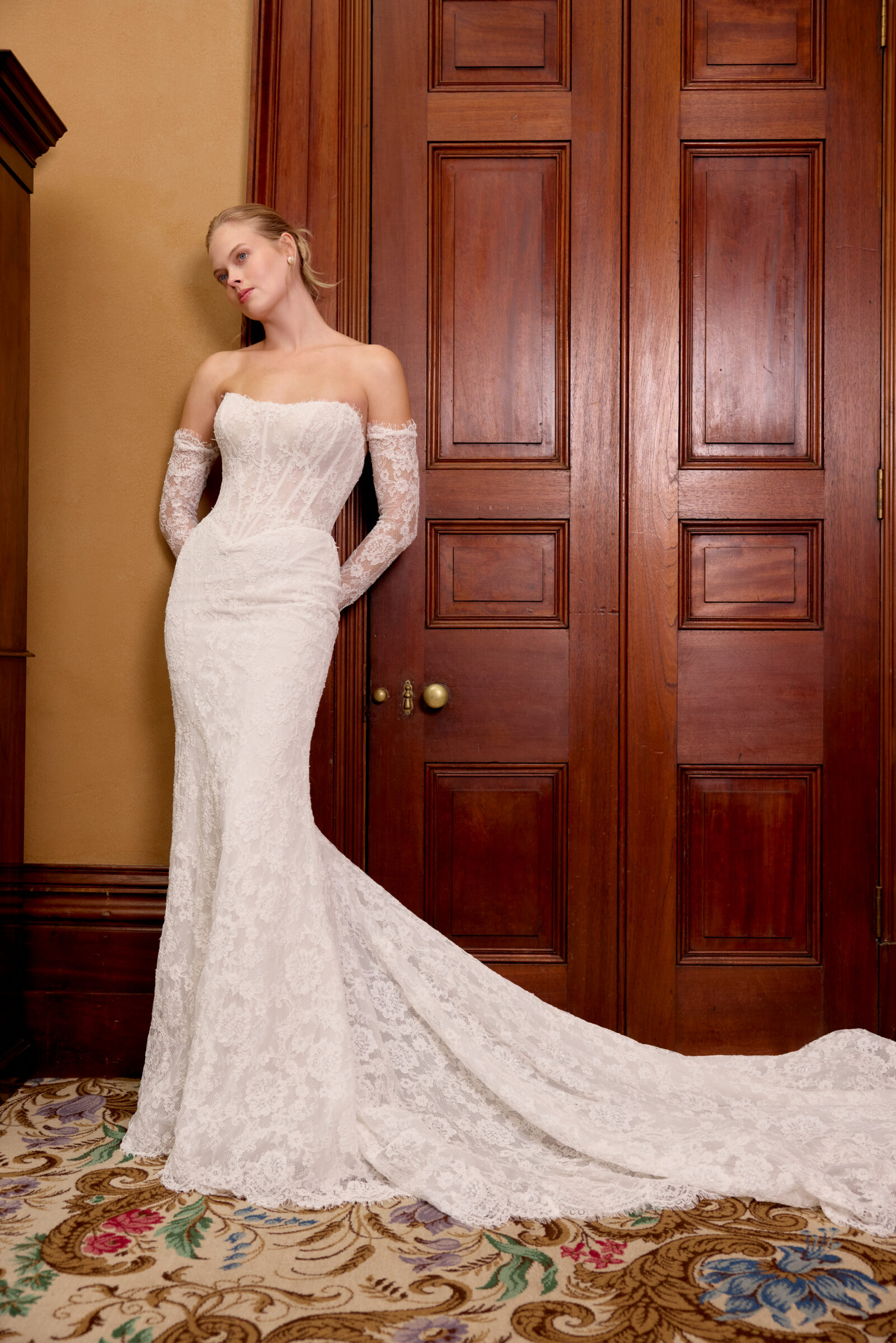 Bride in an off-the-shoulder fitted lace wedding gown with long sheer sleeves and a flowing train, posing against wooden doors.