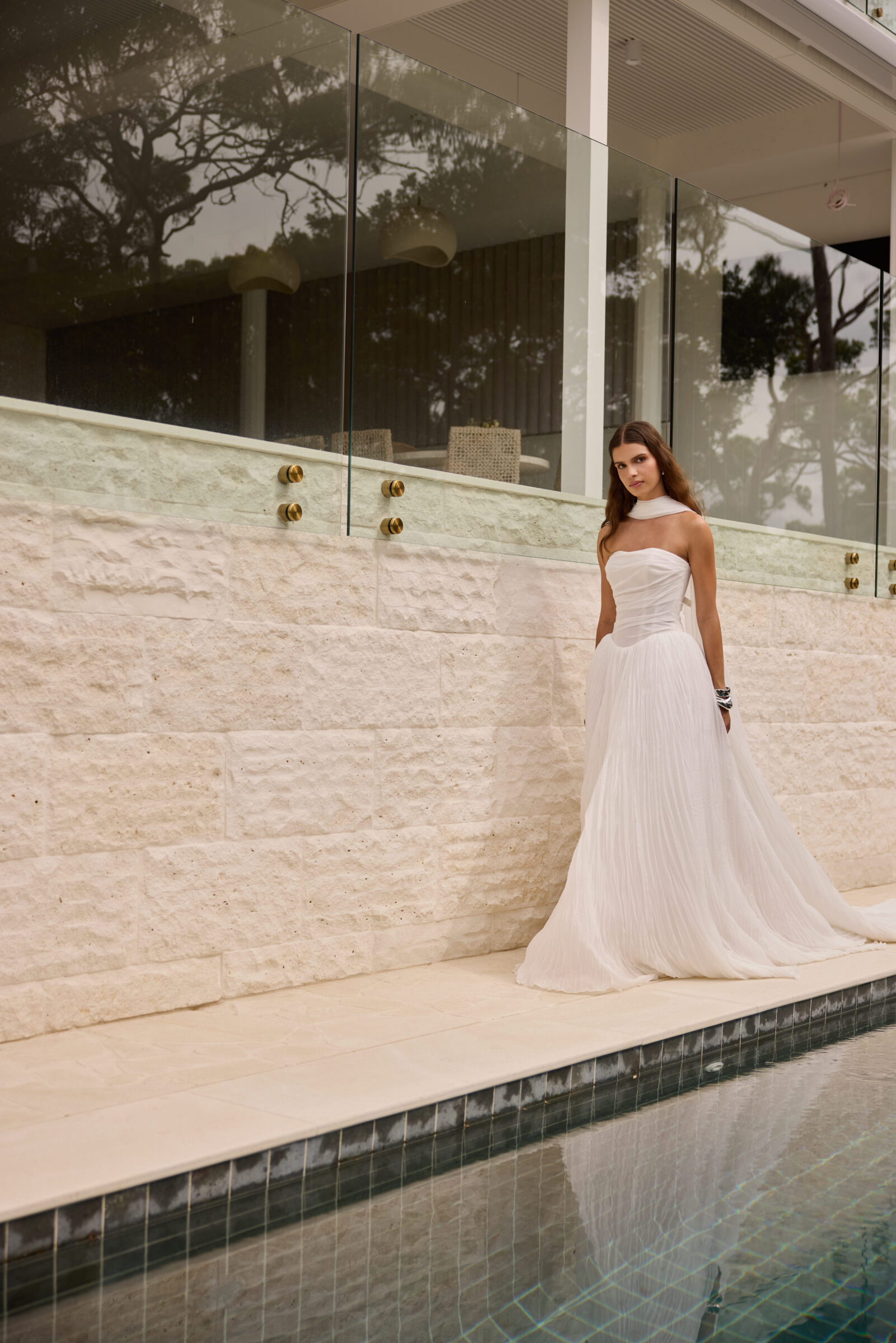 Bride wearing a strapless white wedding gown standing beside a modern pool and stone wall.