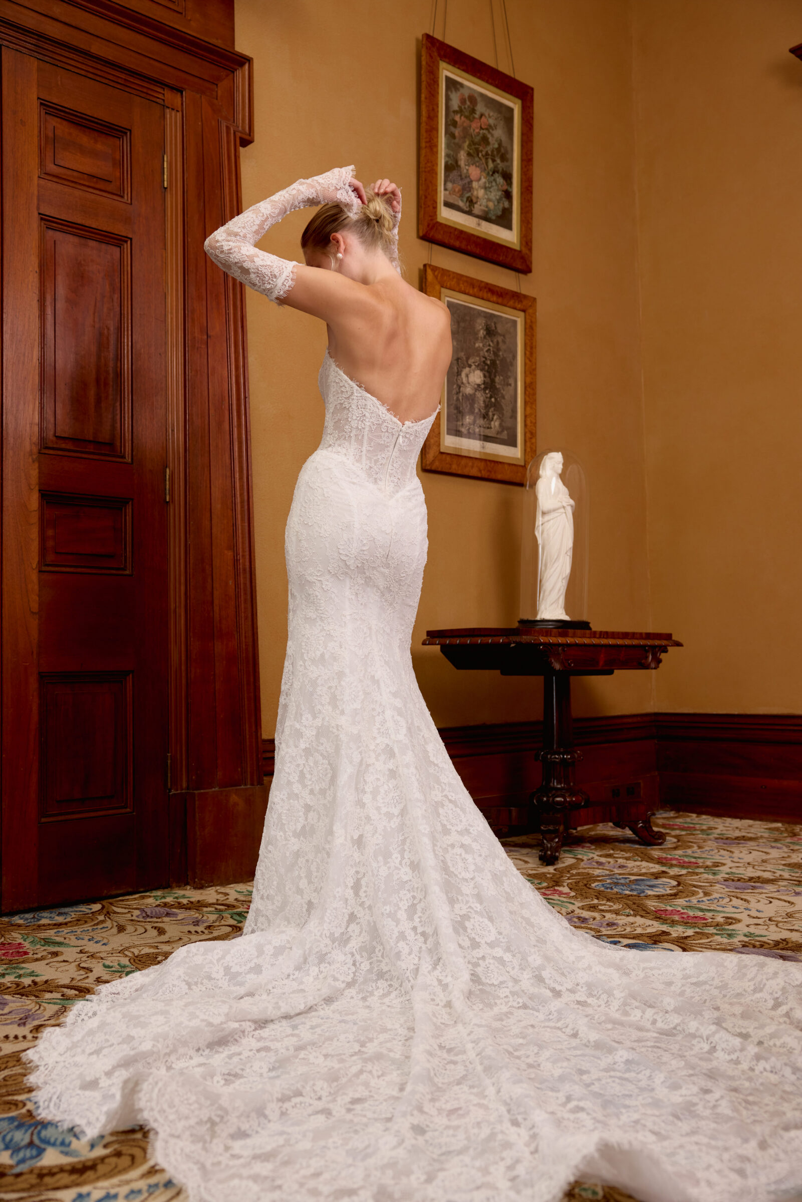 Bride wearing a fitted lace wedding gown with long sheer sleeves and a long train, adjusting her hair in an elegant room.