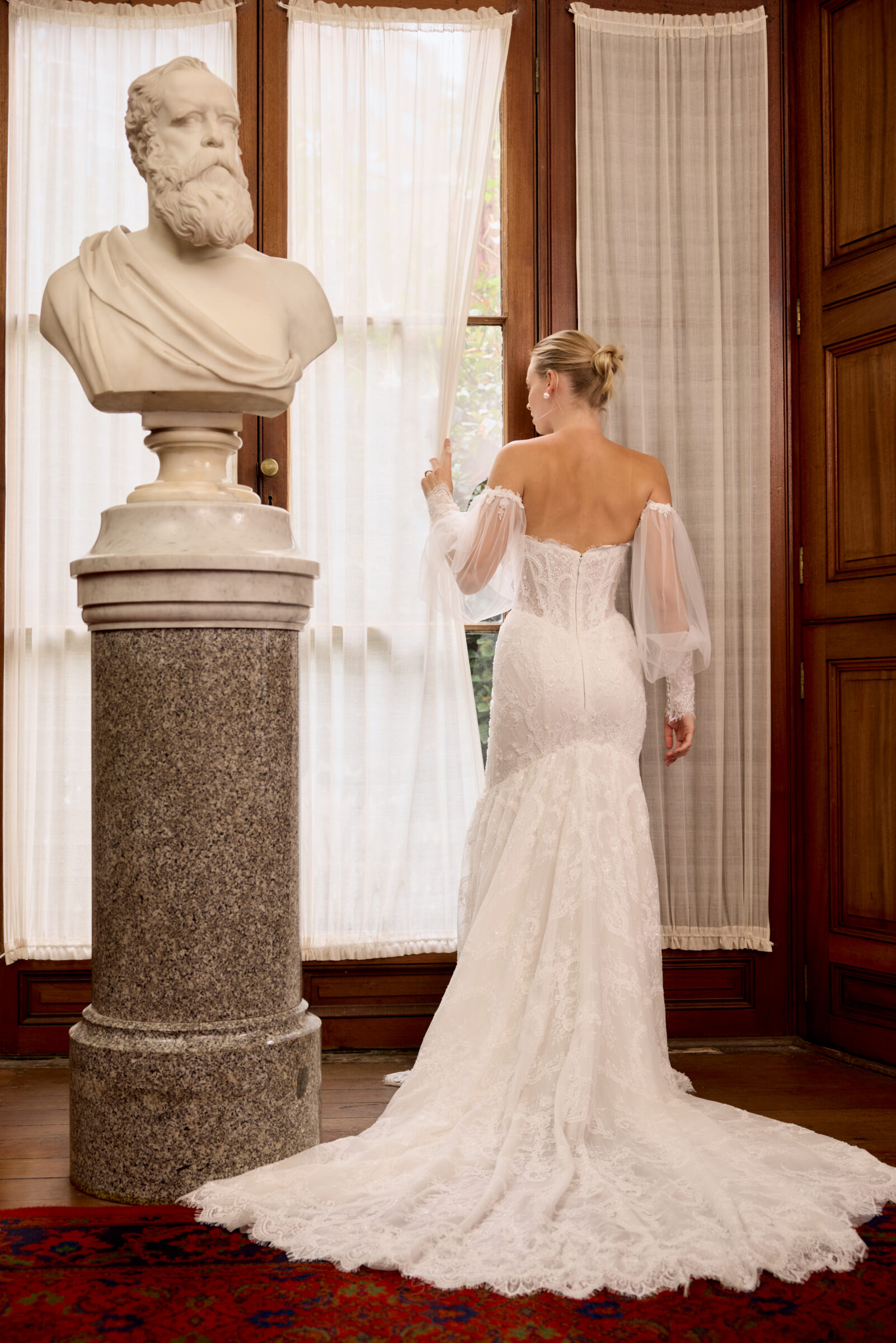 Bride shown from the back in a fitted lace wedding gown with sheer sleeves and a long train, standing beside a classical statue.