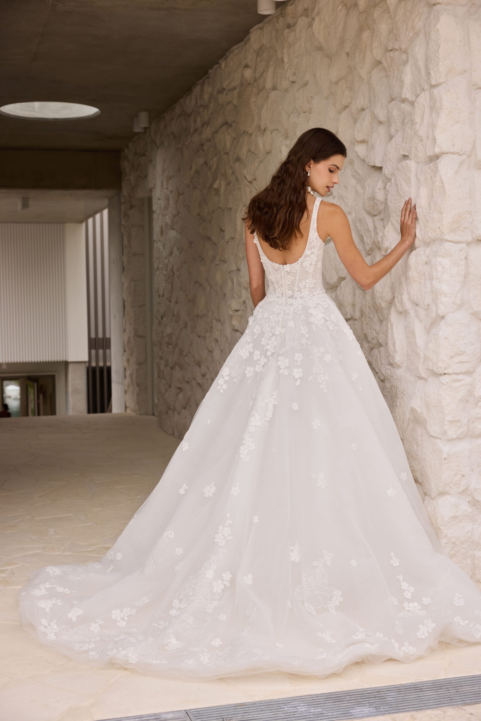 Bride wearing a white lace wedding gown with a full skirt and long train, posing beside a stone wall.