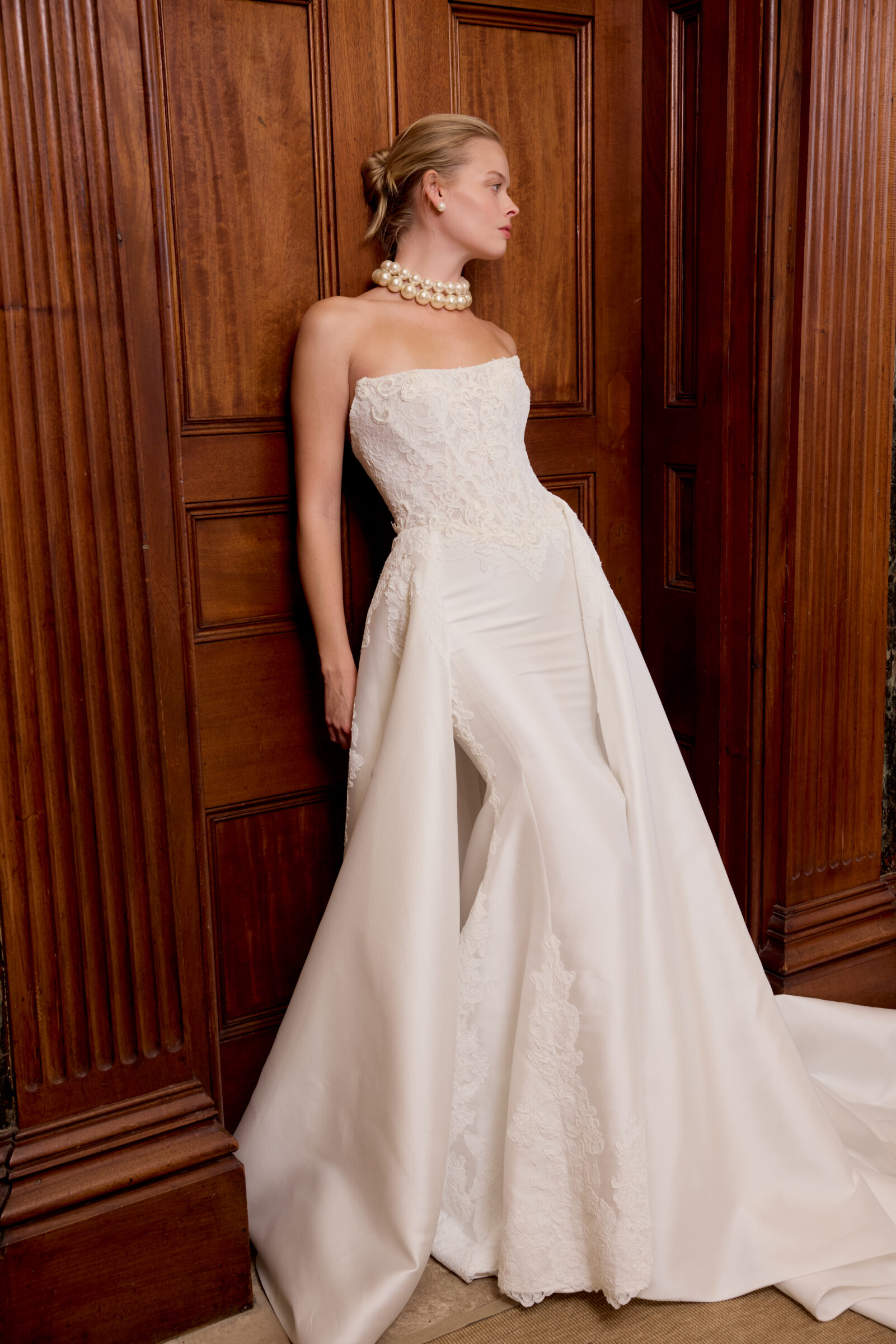 Bride wearing a strapless white wedding gown with a fitted lace bodice, flowing satin skirt, and long train, posing against wooden paneling.