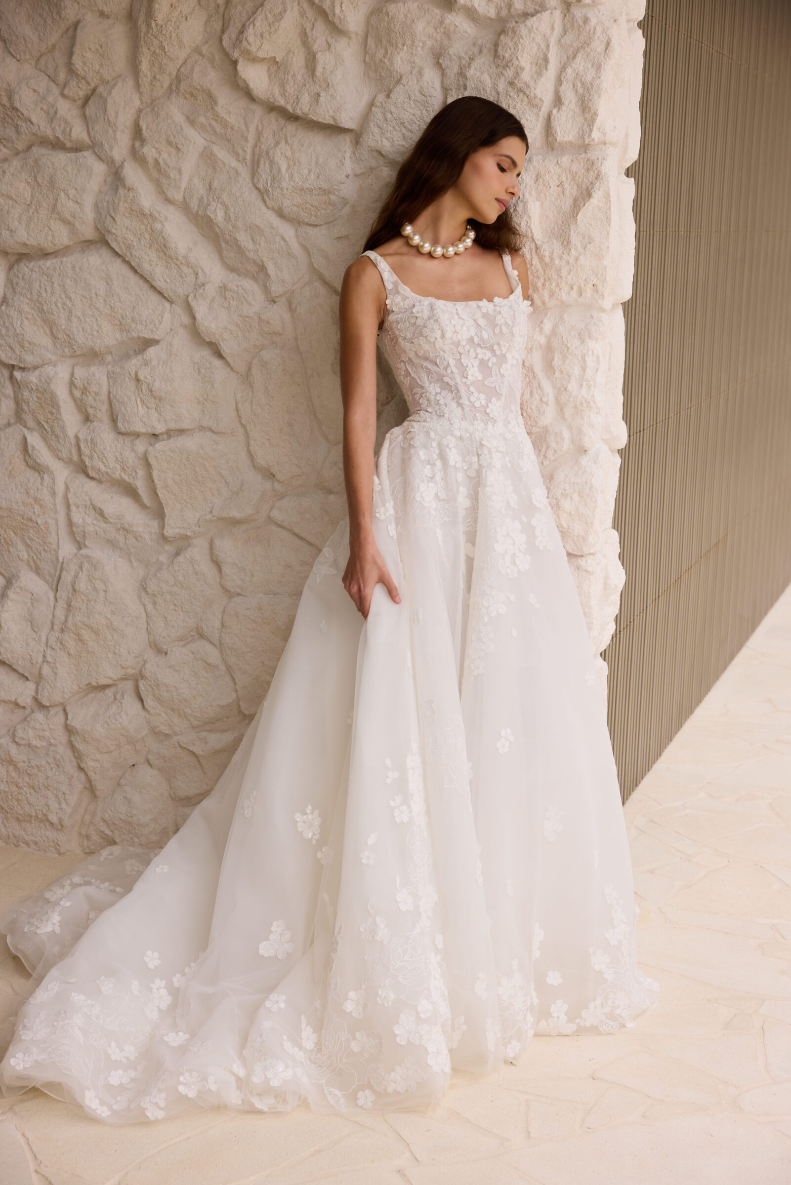 Bride in a white lace wedding gown with a flowing train and sheer neckline, standing beside a textured stone wall.