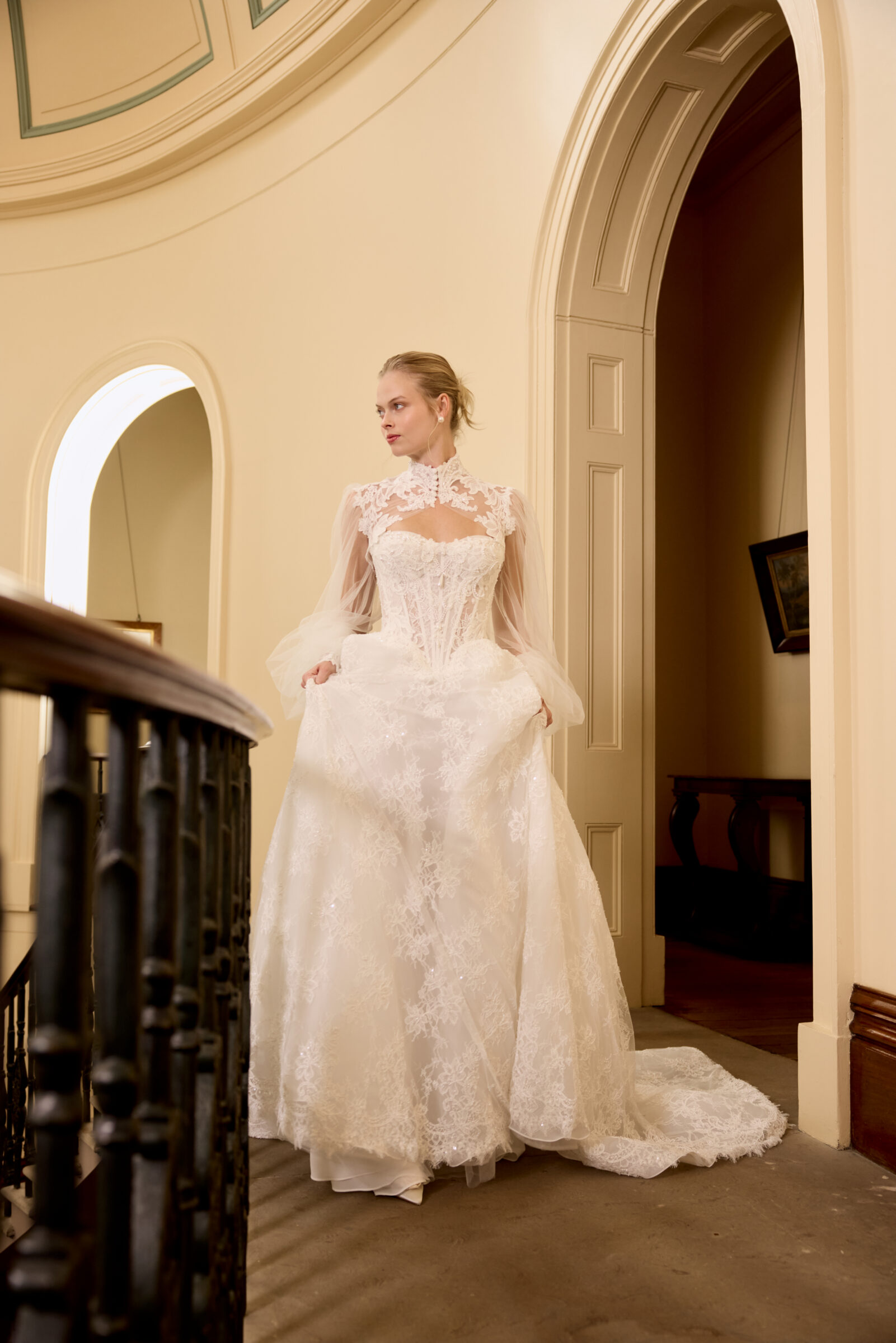 Bride wearing a lace wedding gown with sheer long sleeves and a full skirt, standing in an elegant arched hallway.