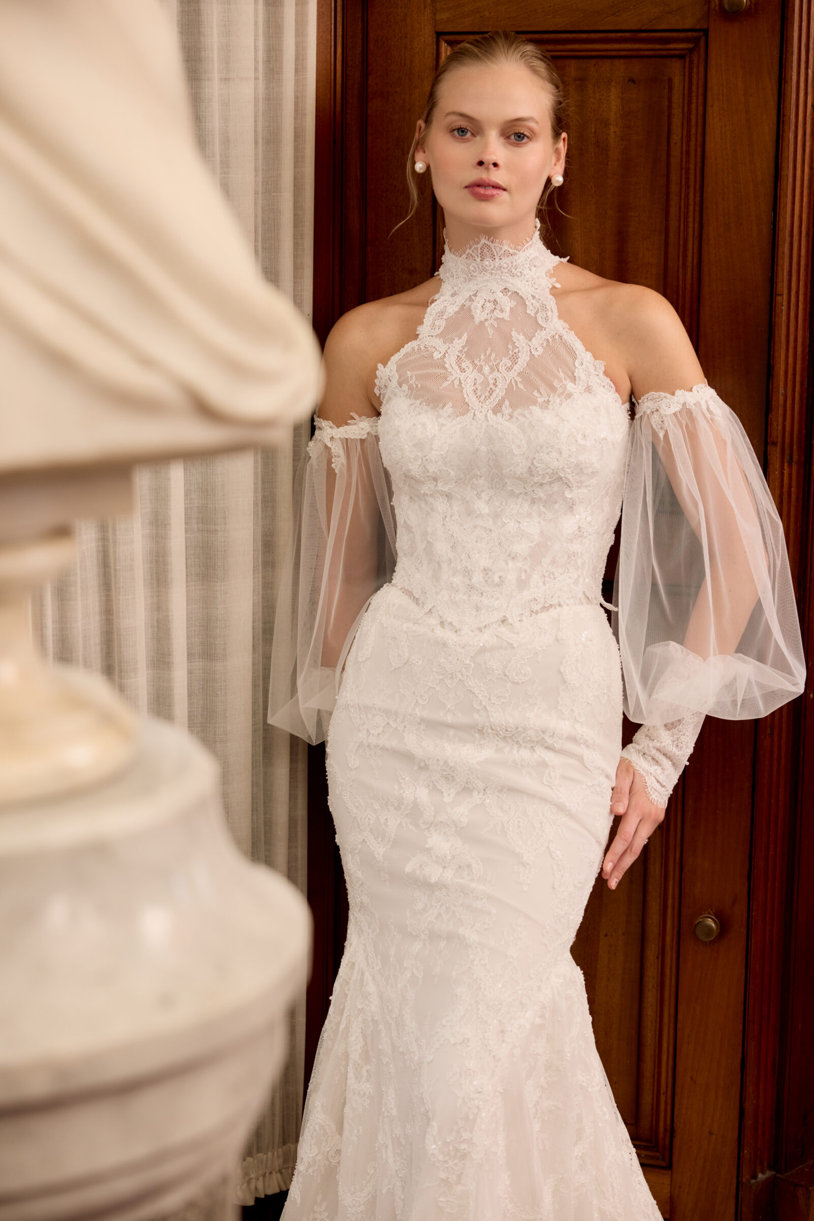 Bride wearing a fitted lace wedding gown with a high neckline, sheer off-the-shoulder sleeves, and pearl jewelry, standing beside a wooden door.