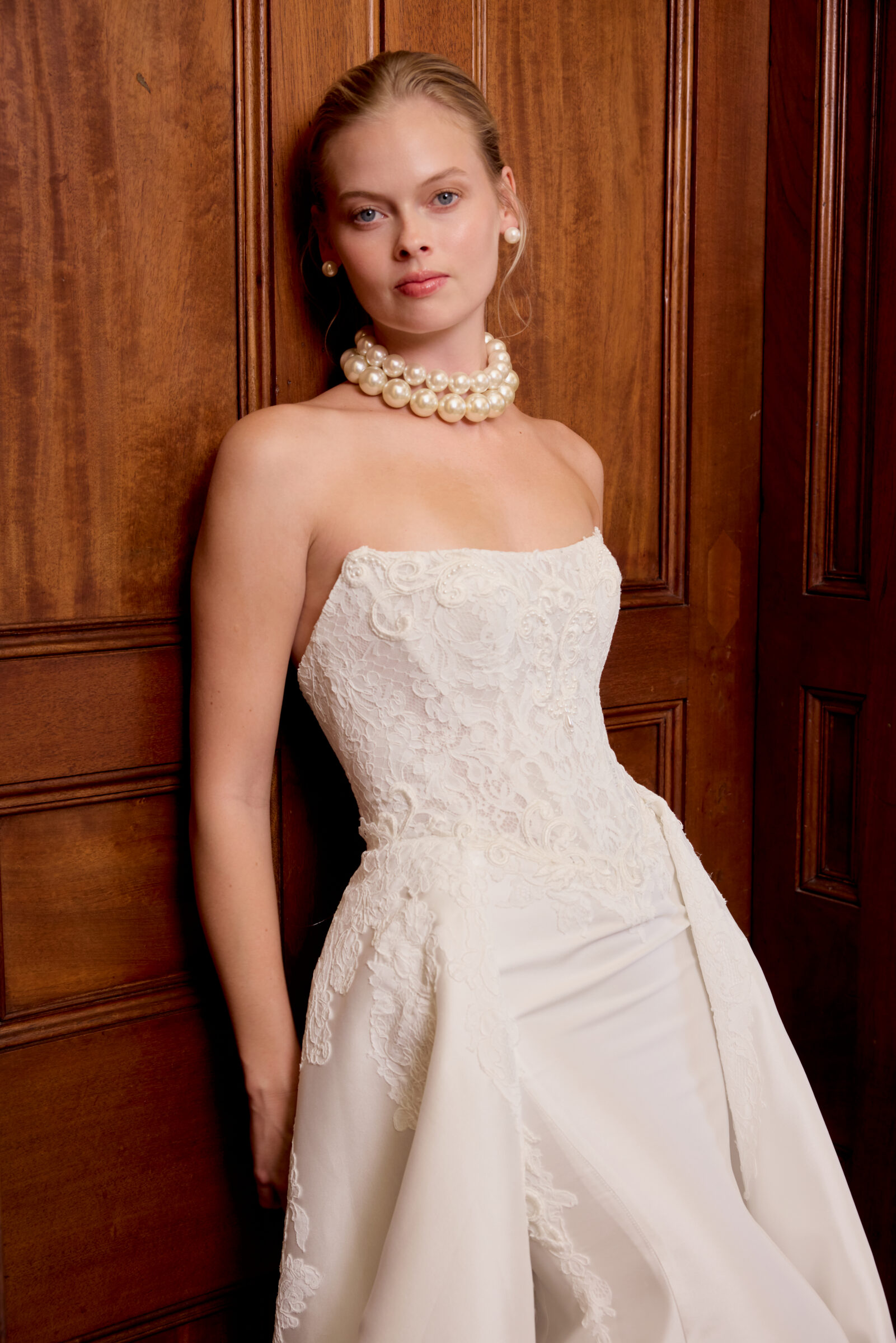 Bride wearing a strapless white wedding gown with a fitted lace bodice and flowing satin skirt, posing against wooden paneling.