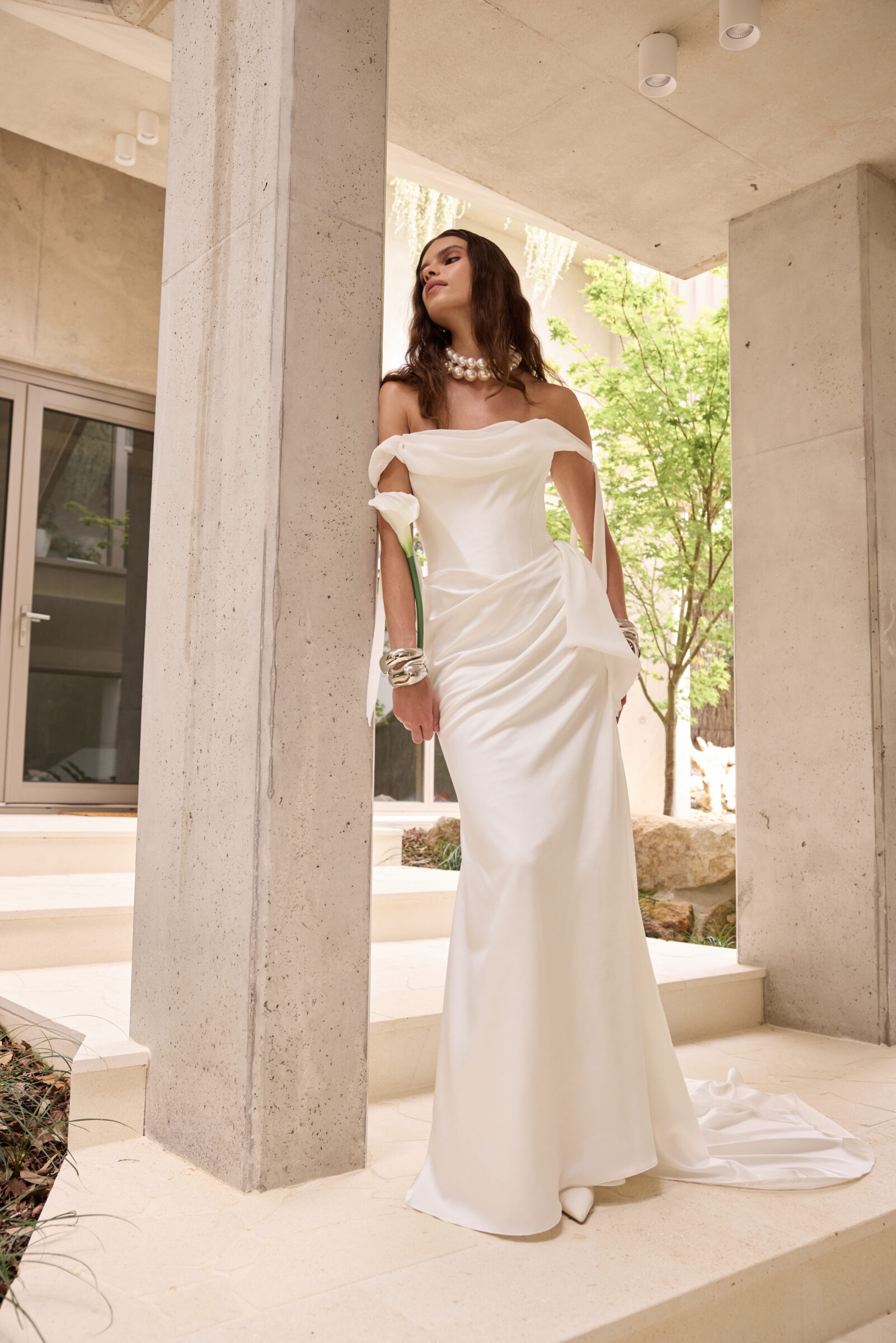 Bride wearing an off-the-shoulder white satin wedding gown, standing on a covered outdoor terrace.