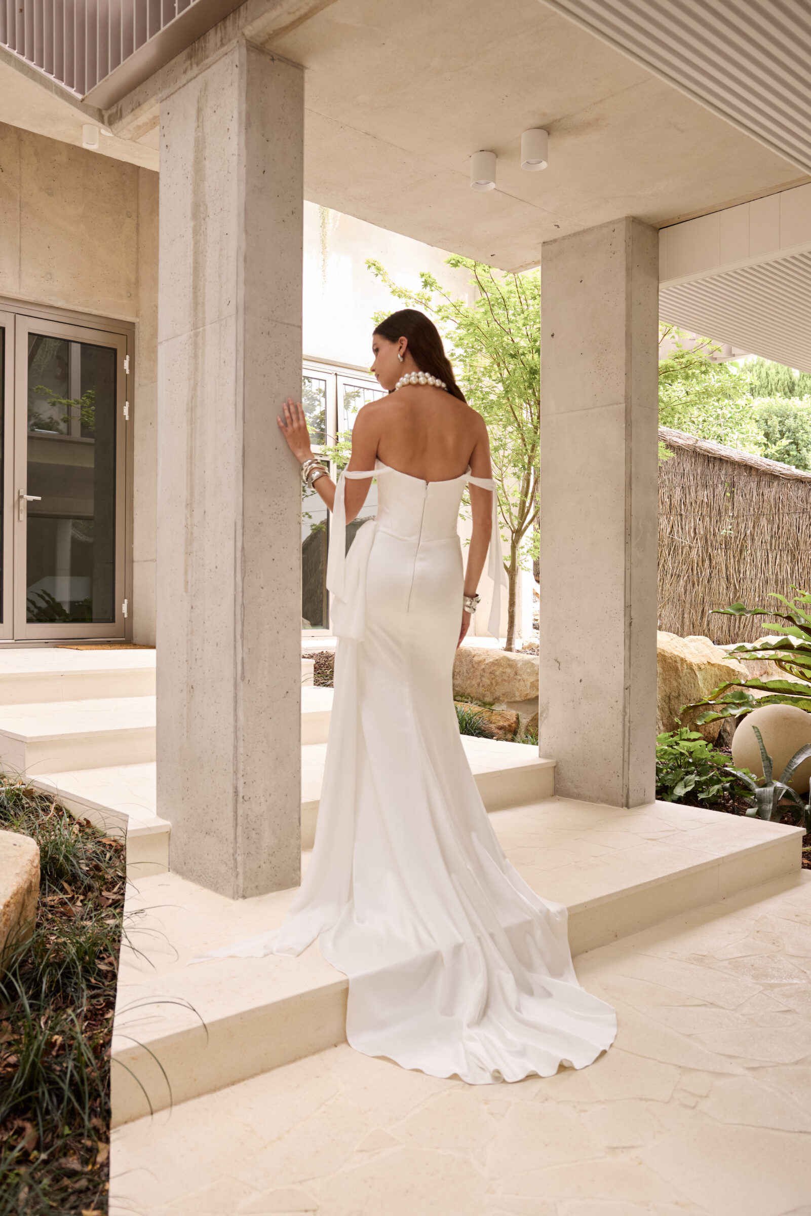 Bride wearing an off-the-shoulder white satin wedding gown with a long flowing train, standing on an outdoor terrace.