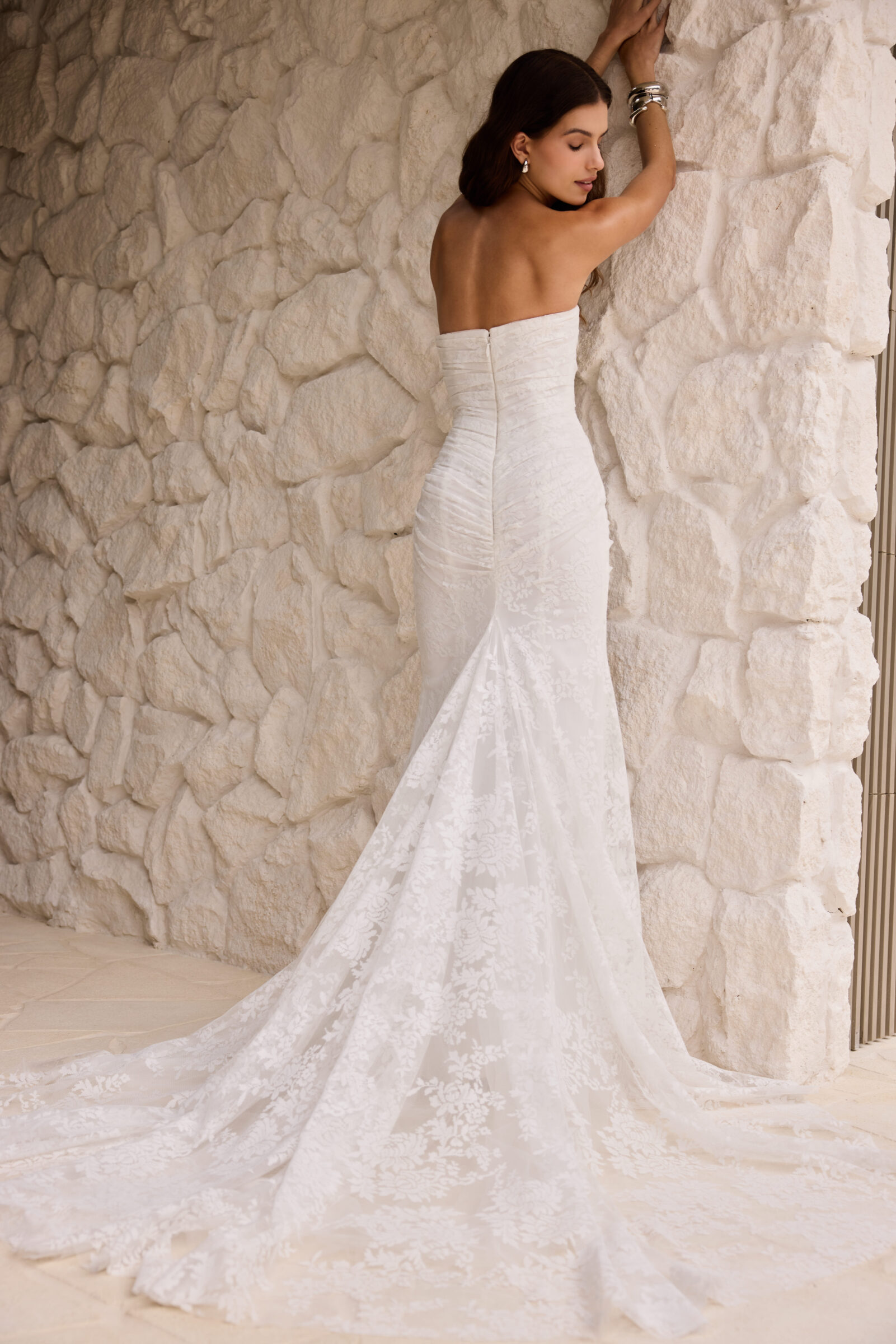 Bride in a strapless fitted white wedding gown with a long train, standing beside a textured stone wall.