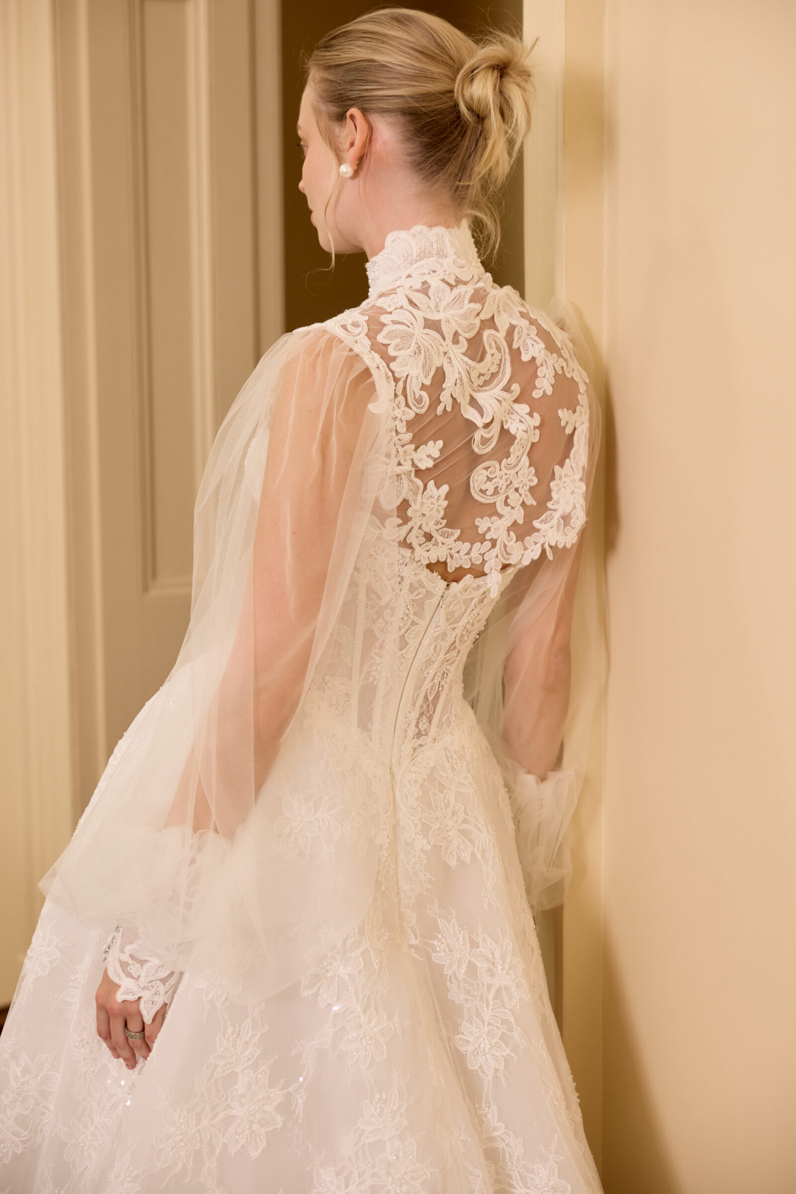Close-up of a bride from the back, showing the intricate floral lace detailing and sheer sleeves of her wedding gown.