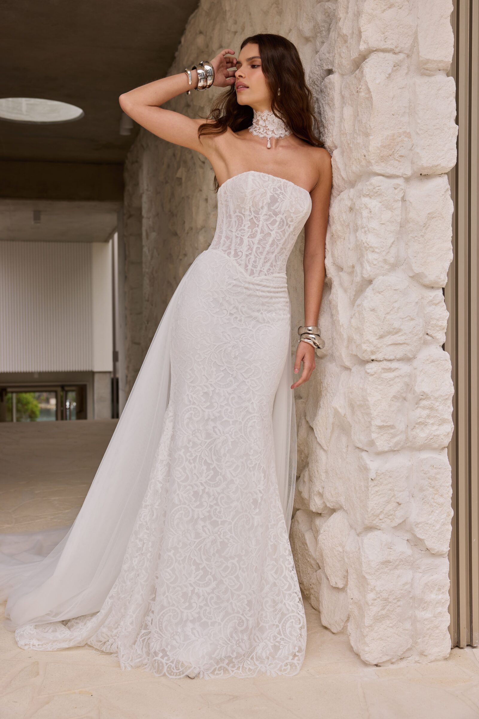 Bride in a strapless white lace wedding gown with a long train, posing beside a stone wall.