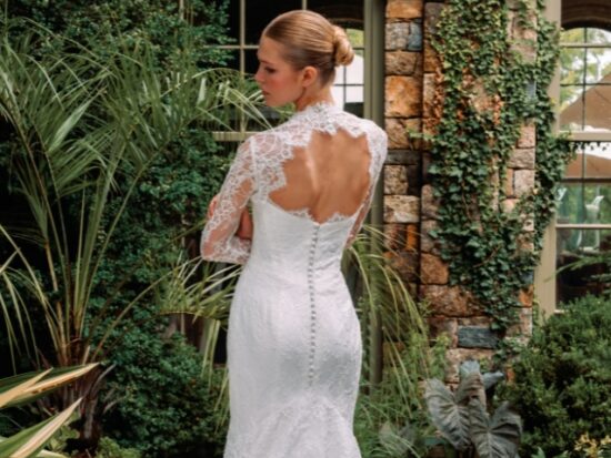 A model poses from behind in a long lace wedding gown with an open back, sheer sleeves, and a scalloped train surrounded by greenery.
