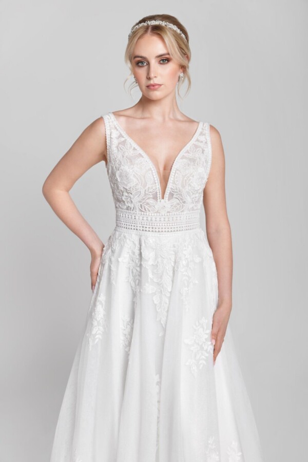 Charming bride to be wearing a wedding dress with a deep v cut neckline, belted bodice and floral detailing on the skirt. This dress also comes with optional lace sleeves.