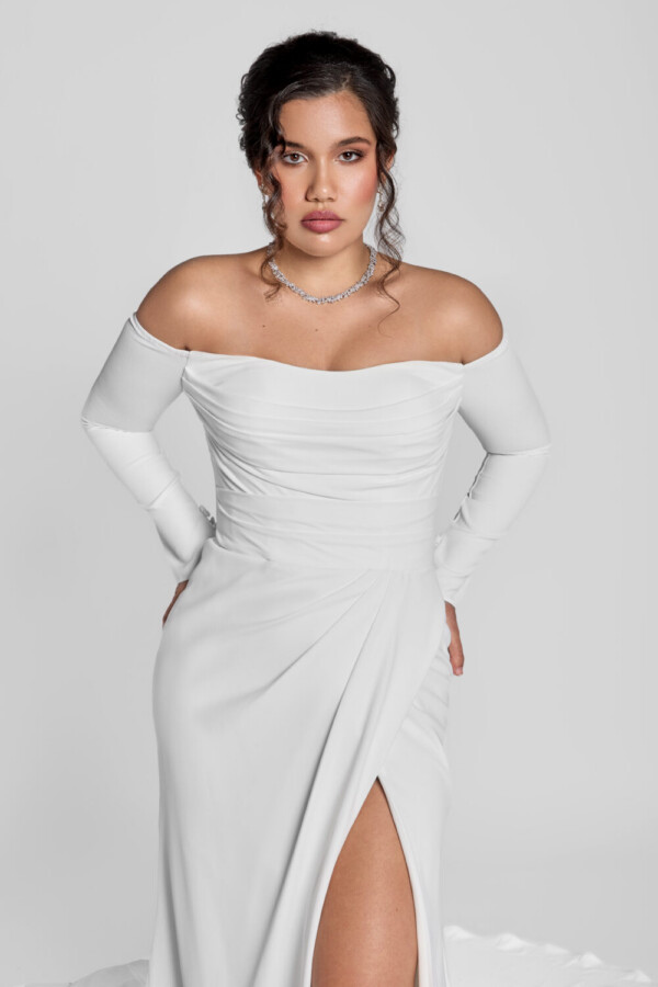 Gorgeous bride wearing a form fitting wedding dress with a ruched bodice and off the shoulder, long sleeves with a high slit in one thigh.