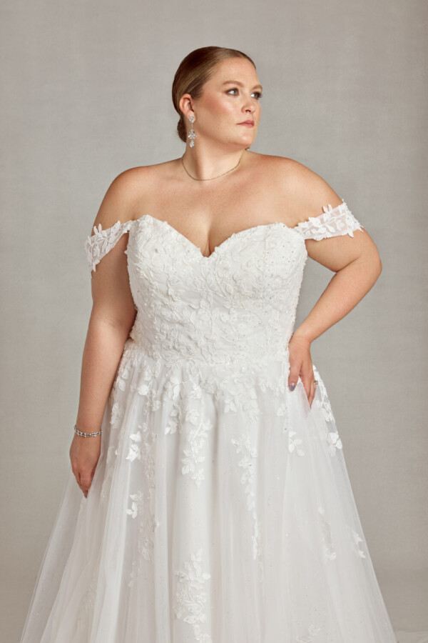 Plus size model wearing her wedding dress with tulle skirt and floral detailing on the bodice and off the shoulder sleeves.